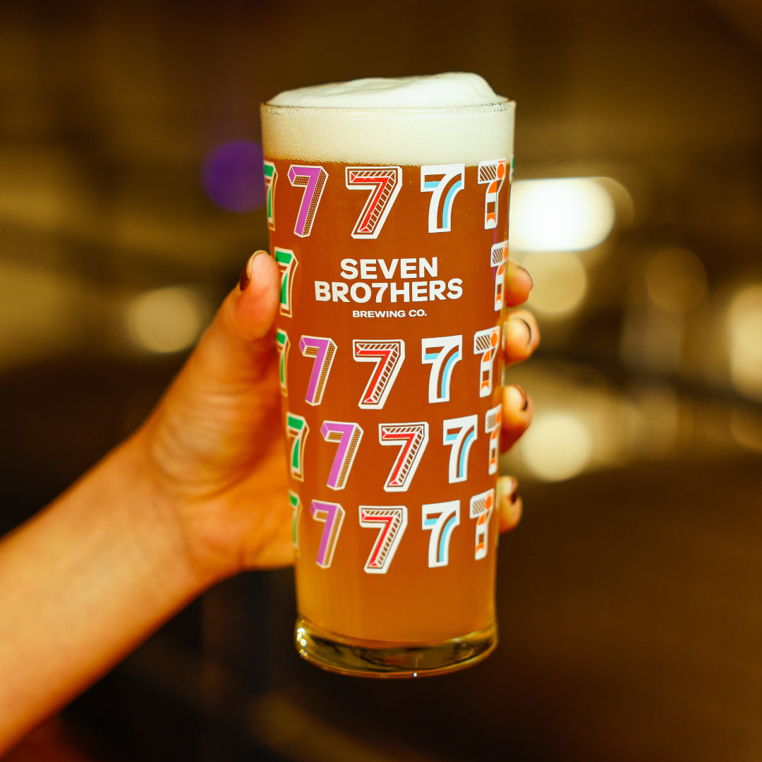 Glass of beer with Seven Brothers Brewing Co. logo held by a hand against a blurred background