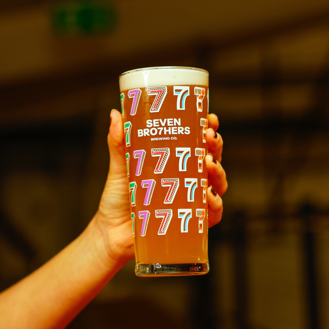Hand holding a glass of beer with 'Seven Bro7hers' branding against a blurred background